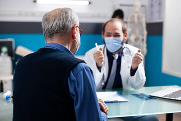 Caucasian doctor sits across retired sick man offering healthcare treatment advice in hospital. Rearview closeup of male pensioner patient discussing with masked physician about his medical concerns.