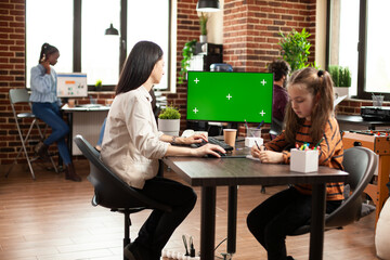 Caucasian female employee using desktop monitor with blank mockup template in brick wall room. Mother and daughter sitting at table with computer displaying isolated greenscreen in startup office.