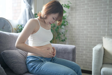 Wall Mural - A pregnant Japanese woman in her 20s wearing clothes that show her stomach is sitting on a sofa in her apartment, holding her big belly.