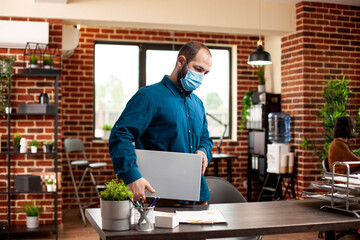 Wall Mural - Bearded man wearing face mask walks into modern office, carrying laptop. Male manager dressed in business casual, stands at desk and appears ready to start busy day in startup company.