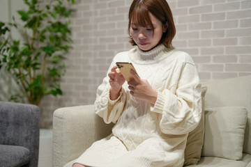 Wall Mural - A pregnant Japanese woman in her 20s wearing a knitted sweater is sitting on a sofa in her apartment and operating a smartphone.