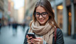 © Vadym - Young woman happily smiles using mobile phone outdoors in city. Looks at smartphone screen intently walking. Woman wears eyeglasses, scarf, jacket. Soft shadows, natural lighting highlight cheerful