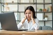 © Vadym - Businesswoman sits at modern office desk looking stressed. Holds head with both hands. Working on laptop. Stressed professional feels overworked. Workplace modern. Young adult. Scene shows tech