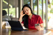 © Wasana - A woman in a red shirt is sitting at a table with a laptop and a cell phone