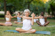 © JackF - Yoga mature female coach conduct rehearsal for multinational followers in park, leading of performing exercises, do fulfil padmasana, svastikasana. People practice sport during yoga workout in nature