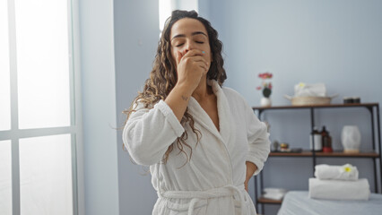 Wall Mural - Woman yawning in a spa room while wearing a white robe, surrounded by a serene interior, showcasing relaxation in a wellness center.