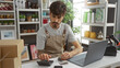 © Krakenimages.com - Young man working in home decor store calculating money with a laptop and calculator, surrounded by home decorations and plants.