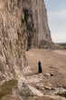 © Milou Dirks - art portrait of woman in long black dress standing on beach near cliffs in Normandy hiding her face