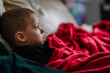 © Raivo - A side profile of a sick child lying on a couch, wrapped in a red blanket, gazing ahead thoughtfully, in a cozy and calm indoor setting.