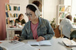 © Mediaphotos - Portrait of Asian young woman wearing headphones while reading book at desk in school or college library focusing on studying for exams copy space