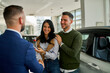 © DusanJelicic - Smiling couple engaging with a car salesman inside a modern showroom. The man and woman appear excited, discussing details about a new vehicle purchase, showcasing a positive buying experience