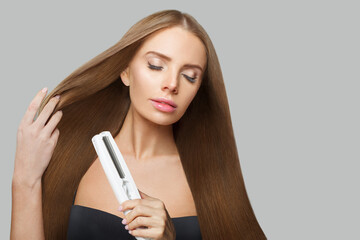 Wall Mural - Glam smiling woman with long straight healthy hairstyle using straightening iron on white background. Beauty, hair care, hair treatment and cosmetic concept