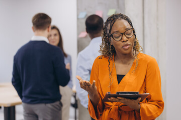 Smiling african american businesswoman holding a folder, diverse business team collaborating, analyzing data on laptops and tablets, sharing insights in contemporary workspace