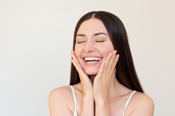 Wall Mural - A young pretty Caucasian brunette woman smiling, closing her eyes and gently touching her face with her hands. 