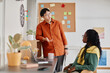 © Seventyfour - Asian man standing while talking to seated Black woman in contemporary office with bulletin board and various office supplies visible in background