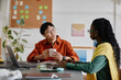 © Seventyfour - Asian man and Black woman engaging in conversation in modern office environment with notes on wall. Both seated at desk with plants in background, exchanging focused ideas