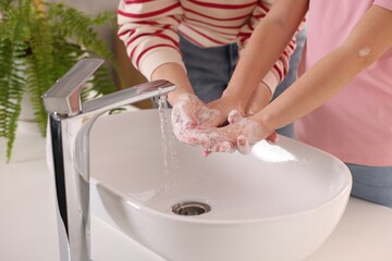 Wall Mural - Mother and daughter washing their hands above sink indoors, closeup