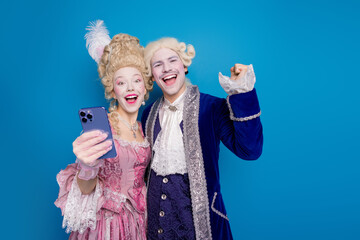 Wall Mural - Elegant couple in baroque costumes capturing joy with a smartphone against a vivid blue background.