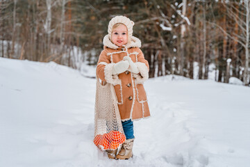 Child girl in a beige coat or in winter sheepskin coats and hats in a snowy forest. A three-year-old girl has fun in winter. Kid in winter Russian folk clothes, like in a fairy tale. One person 