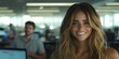 © Imagine Crane - A young woman with long hair smiles confidently in a modern office filled with computers. Her colleague is seen focused on work in the background as natural light illuminates the space