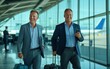 © Premium_art - Two smiling businessmen in suits pull luggage through an airport.