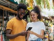 © TPS Studio - A couple enjoys a moment together while looking at a smartphone, surrounded by vibrant market stalls and greenery.