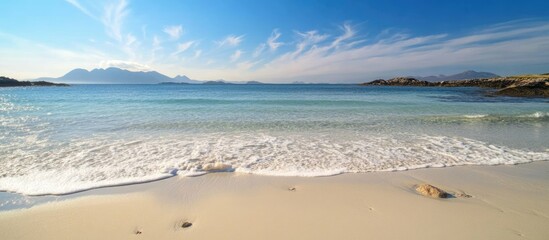 Aufkleber - Tranquil sandy beach with gentle waves and scattered rocks under a clear blue sky during high tide at a picturesque seaside location