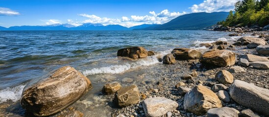 Canvas Print - Rocky shoreline with clear waters and distant mountains under a blue sky, capturing a serene natural landscape by the lake.