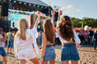 © artiemedvedev - Back view of three women enjoying a music festival on sandy shore, raising drinks in cheers under sunny sky. Casual summer outfits, energetic crowd gathered by stage for live performance vibes.
