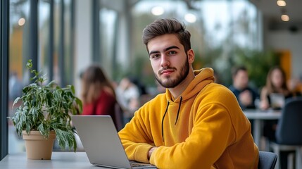 Poster - Young man in yellow hoodie working on laptop in bright modern cafe, urban work or study environment