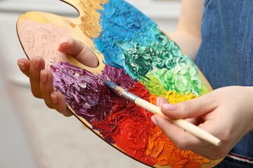 Wall Mural - Woman with paintbrush mixing paints on palette against blurred background, closeup