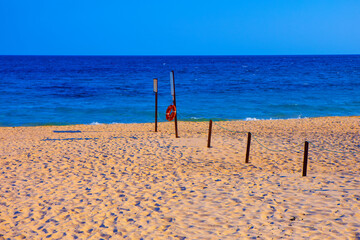 Wall Mural - Sandy beach with a clear blue sky and ocean in the background. There are several poles with signs and a lifebuoy attached to one of them, as well as a rope barrier leading towards the water