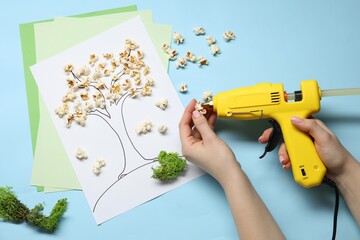 Wall Mural - Woman with hot glue gun making craft on light blue background, top view