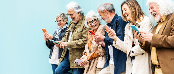 Group of senior people using technology devices together standing on a blue wall - Happy older friends having fun watching funny video on smartphone - Tech and modern elderly concept	