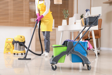 Poster - Trolley with cleaning supplies against female janitor vacuum cleaning floor in office, closeup