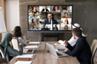 © fizkes - Group of multicultural businesspeople take part in videoconference, sit at conference table looking at TV screen displayed diverse business partners, engaging in virtual meeting, negotiating remotely
