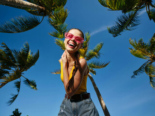 Poster - Woman in sunglasses and yellow top stands before palm trees under clear blue sky, epitomizing summer, relaxation, and tropical vibes