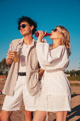 Young stylish friends posing at the seaside. Fashion man and cute female dressed in casual summer clothes. Smiling models having fun. Cheerful women and guy outdoors, Hold and drink lemonade