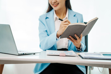 An Asian female accountant works happily at her desk in the office, using a laptop to analyze financial charts. She manages business finance and online transactions for company success