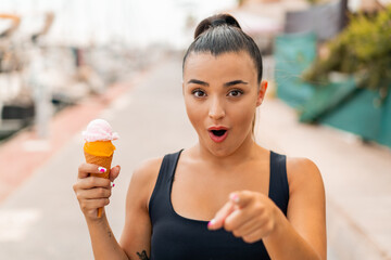 Wall Mural - Young pretty woman with a cornet ice cream at outdoors surprised and pointing front