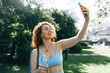 © shurkin_son - Redhead fitness blogger recording video or stories, holding smartphone and taking selfie to share with her followers walking in park after training workout outdoors with bottle of water in hand