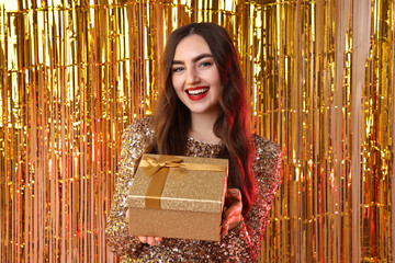 Happy young woman with gift box against golden foil curtain