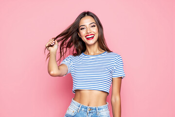 Wall Mural - Cheerful young woman in striped crop top smiling and playing with her hair against pink background.