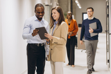 Wall Mural - Business colleagues using a digital tablet while walking through a busy office corridor, discussing work related matters