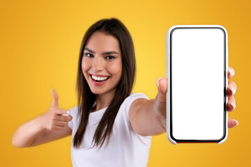 Aufkleber - Cheerful young lady holding smartphone with white empty screen, pointing at blank display for mobile application advertisement or your text, gadget mockup template, orange studio background