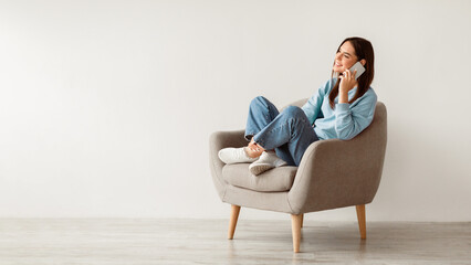 Poster - Friendly millennial woman speaking on mobile phone, sitting in armchair, enjoyng conversation with friend against white wall, banner with empty space. Modern communication concept