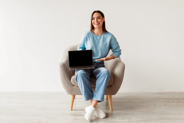 Poster - Check out this new website. Happy young lady sitting in armchair with laptop computer, showing blank screen with mockup, advertising your site design, full length portrait