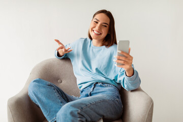 Poster - Cheerful young woman having online business meeting on smartphone, sitting in cozy armchair on white studio background. Millennial student taking part in webinar, communicating remotely on webcam