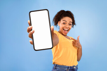 Poster - Cheery millennial African American woman pointing finger at smartphone on blue studio background, mockup for mobile app on white screen. Cellphone website template, online store advertisement