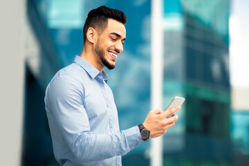 Wall Mural - Positive young handsome arabic businessman with mobile phone outdoors, happy middle-eastern entrepreneur reading emails on cellphone, enjoying success, business building background, copy space